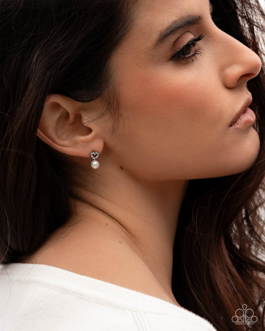 A close-up side profile of a woman wearing a silver stud earring with a dangling white pearl, set against her dark hair and white top.
