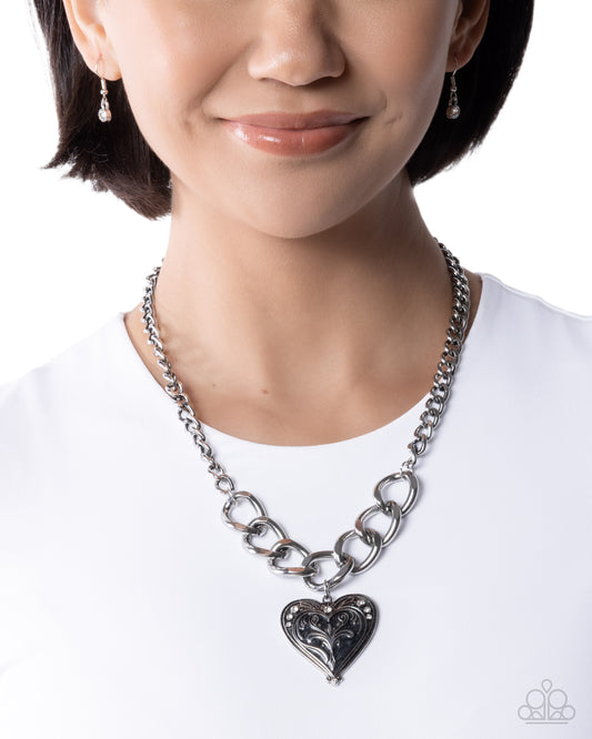 Person wearing a silver chain necklace with a heart-shaped pendant on a white background