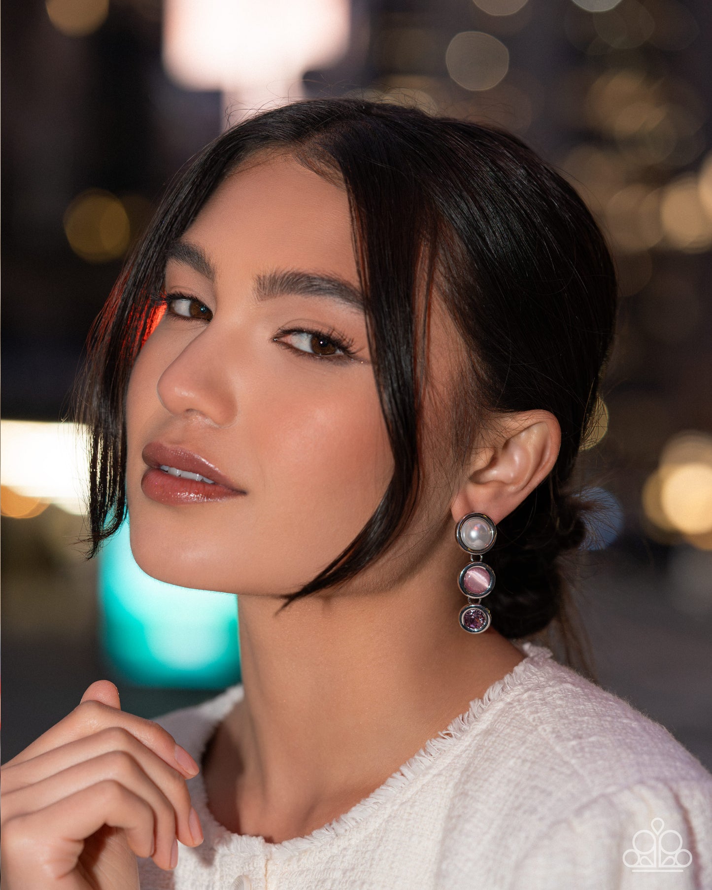 A close-up portrait of a woman wearing a dangling pearl and pink gemstone earring, with a softly blurred bokeh background.
