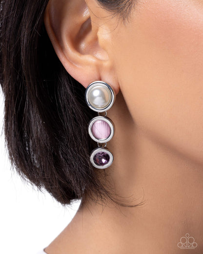 A close-up of a model's ear wearing a silver drop earring with three circular charms: a white pearl, a pink textured stone, and a dark purple stone.