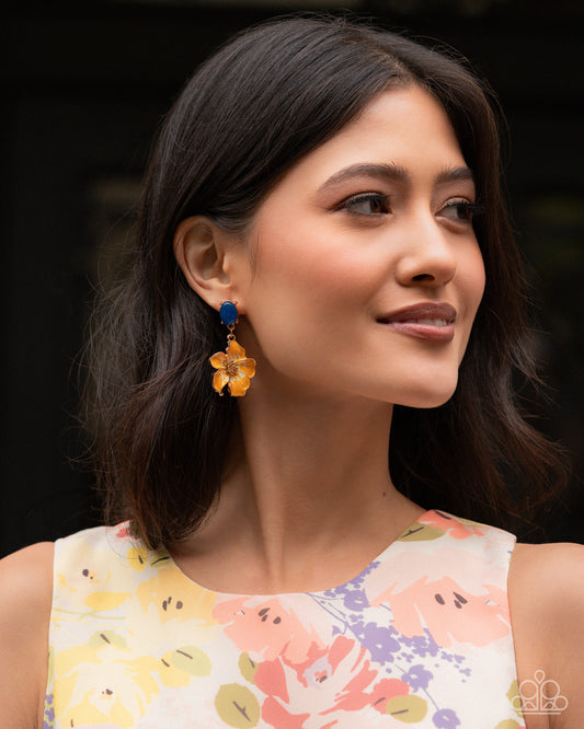 A woman with dark shoulder-length hair wearing a sleeveless floral top and a dangling flower earring with a blue top and amber-yellow petals, shown in a close-up portrait.