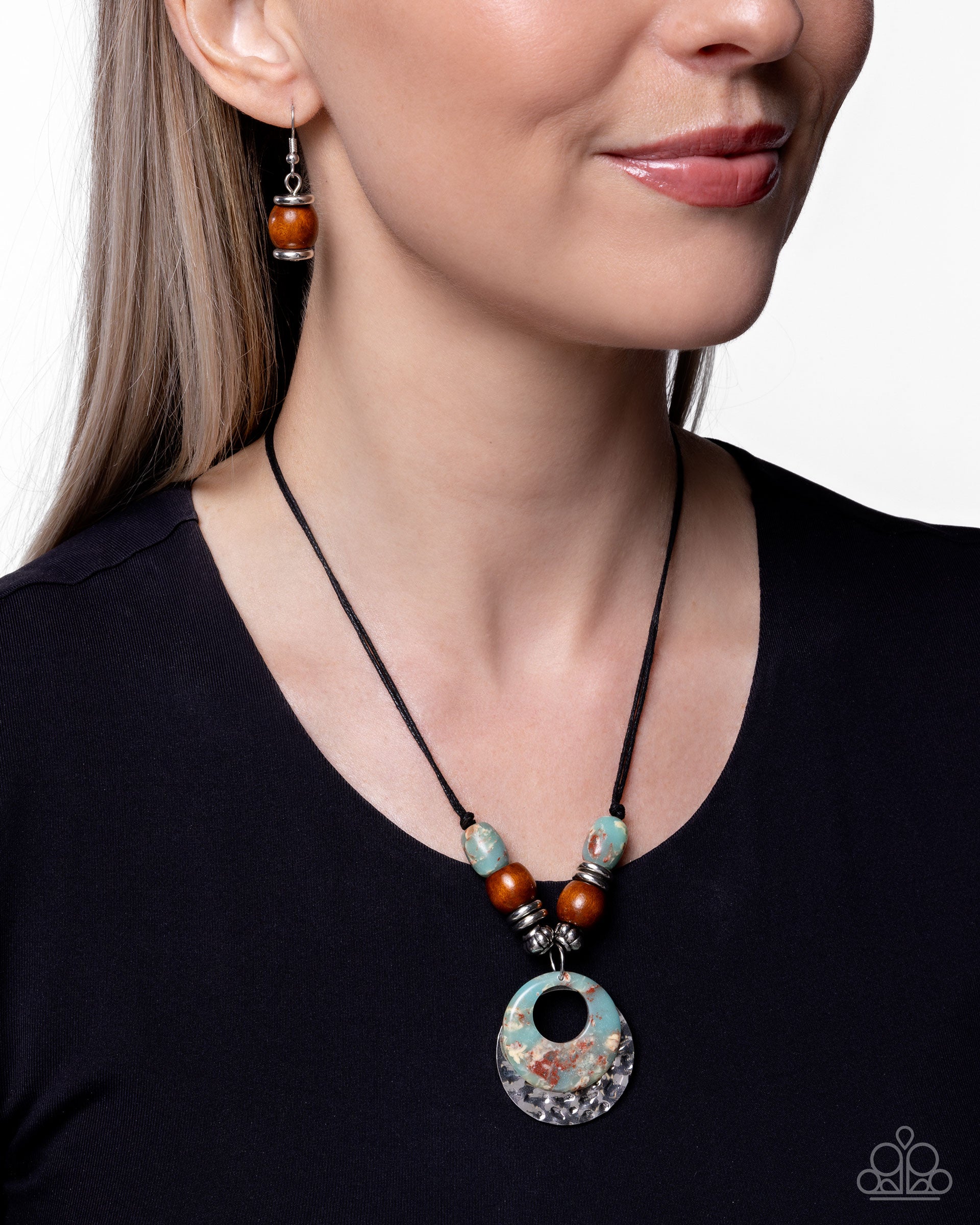 A close-up of a woman wearing a black cord necklace with a circular pendant made of marbled turquoise, brown beads, and silver-toned metal details. She is also wearing a matching drop earring.