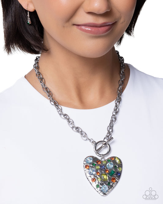A close-up of a model wearing a silver chain necklace with a heart-shaped pendant covered in colorful rhinestones, on a white background.