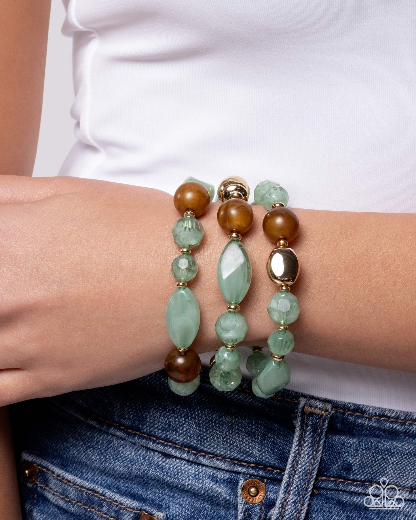 Three beaded stretch bracelets on a model's wrist, featuring light green stone beads, brown beads, and gold-toned metal accents, worn with a white top and blue jeans.