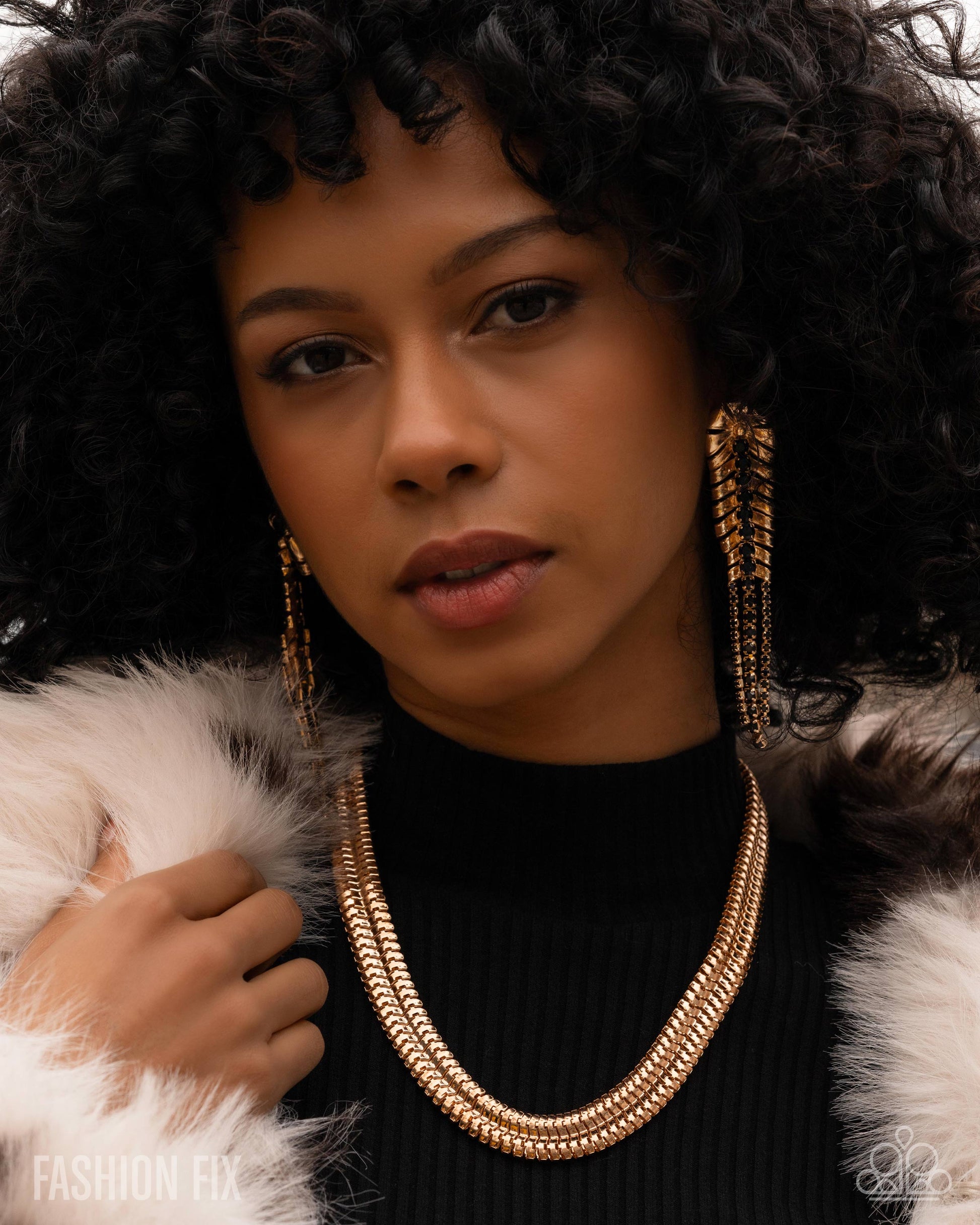 Woman wearing a gold necklace and earrings with a fur coat, posing against a neutral background.
