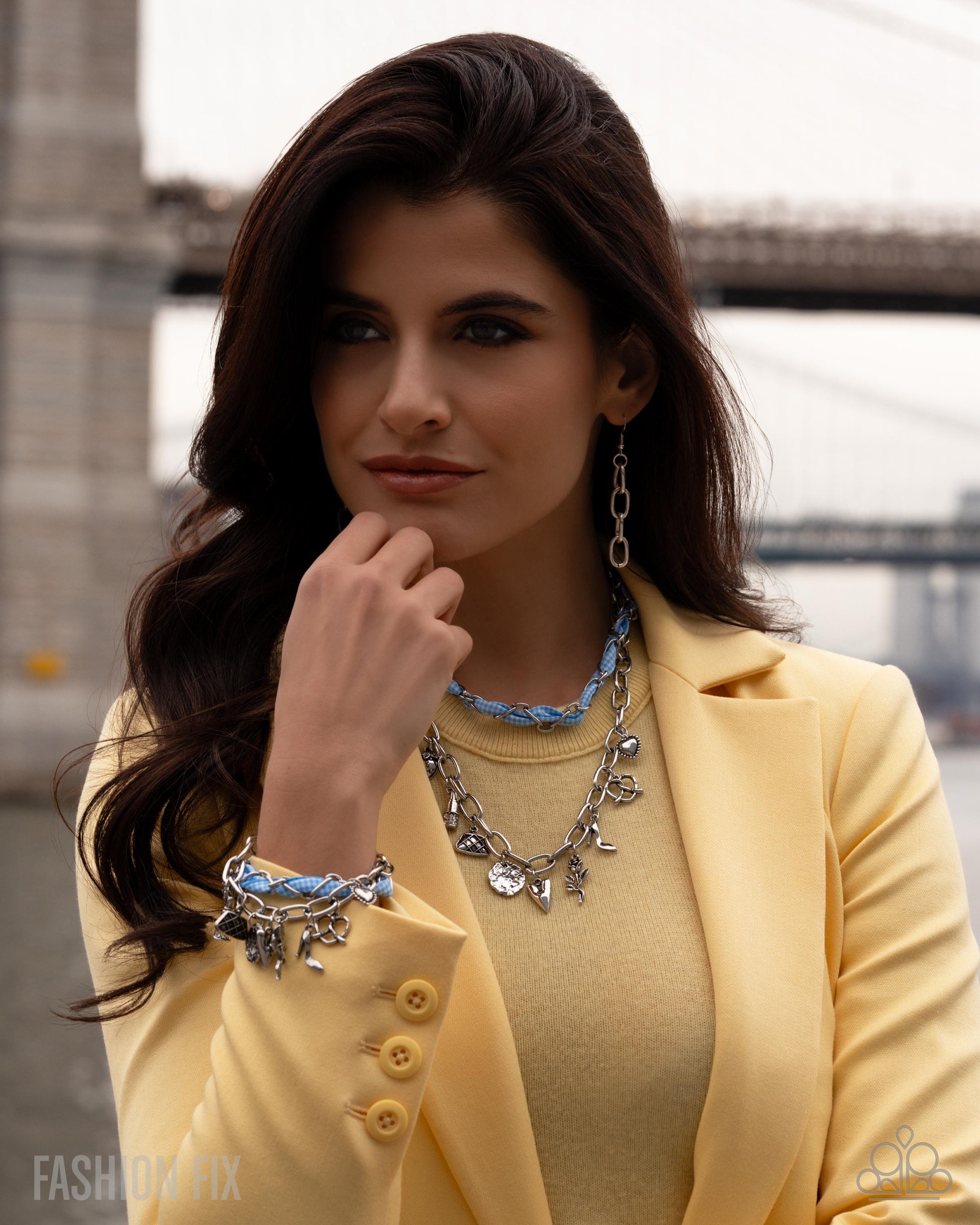Woman wearing a yellow jacket and sweater with a necklace, standing in an urban setting.