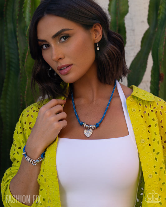 Woman wearing a yellow lace top over a white dress with a blue necklace, standing against a cactus background.