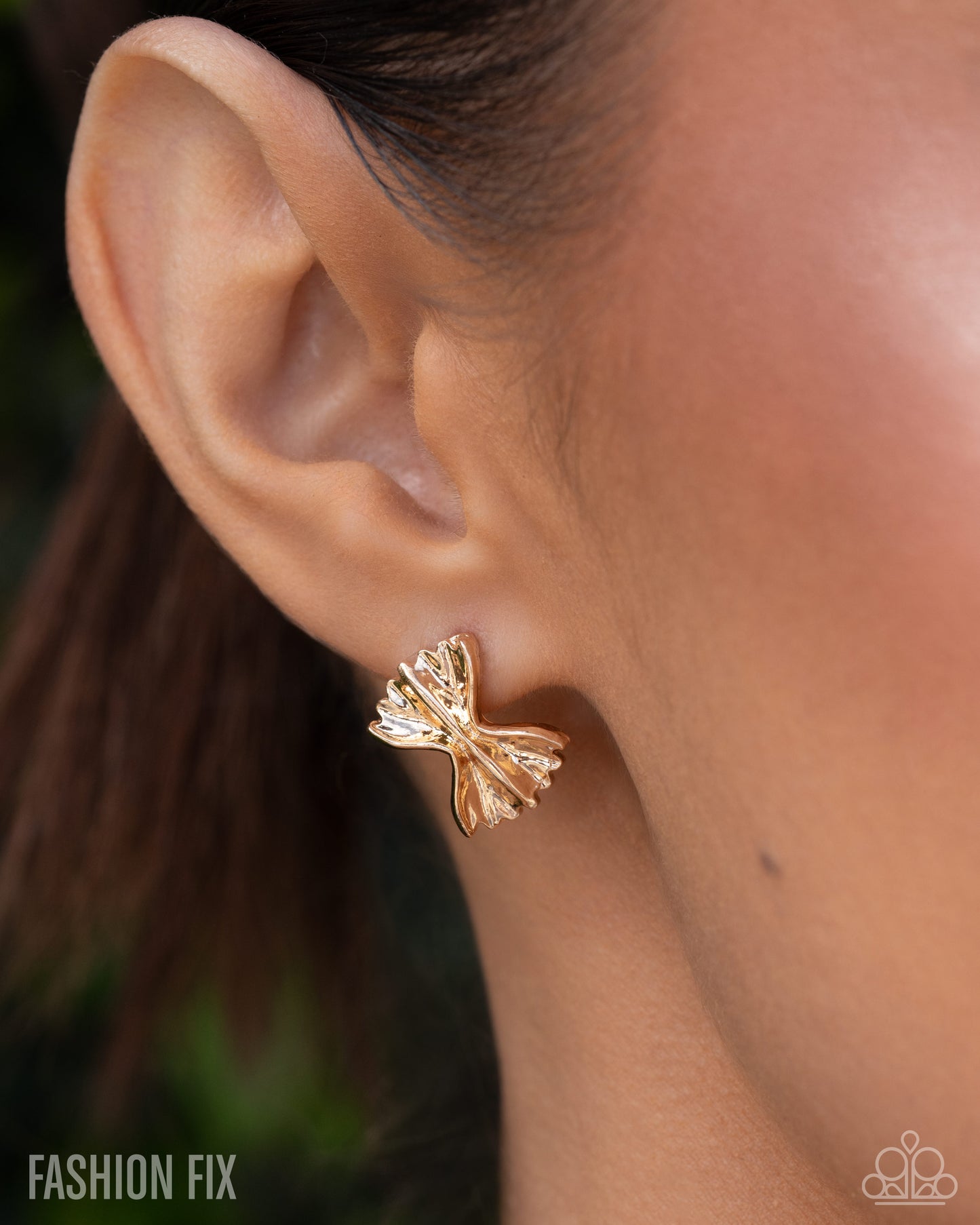 A close-up of a model's ear wearing a small gold bow-shaped stud earring. The image features a 'FASHION FIX' watermark in the bottom-left corner and a small logo watermark in the bottom-right.