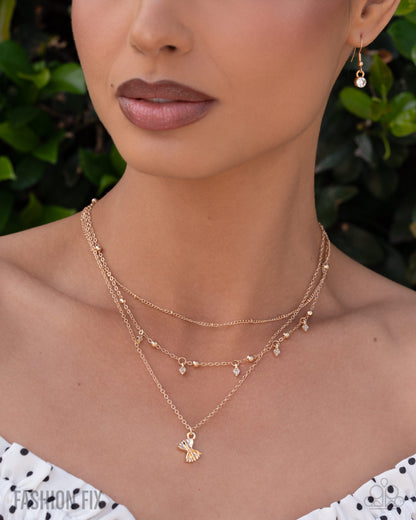 A close-up of a model wearing a layered gold necklace with a small bee pendant, styled with a matching drop earring against a background of green foliage.