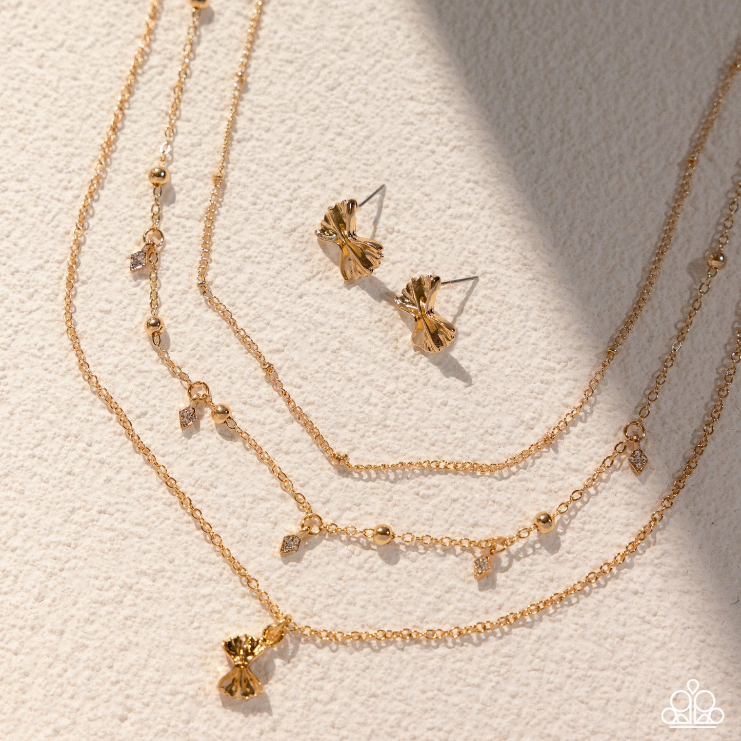 A gold-toned jewelry set arranged on a textured off-white surface, including a layered chain necklace with small dangling charms, a matching stud earring, and a slim bangle with a bow charm. The jewelry is photographed in warm directional light with soft shadows.