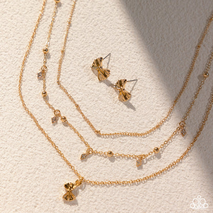 A gold-toned jewelry set arranged on a textured off-white surface, including a layered chain necklace with small dangling charms, a matching stud earring, and a slim bangle with a bow charm. The jewelry is photographed in warm directional light with soft shadows.
