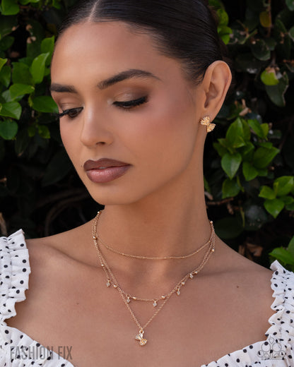 A close-up lifestyle shot of a woman wearing a delicate gold double-layered necklace with butterfly charms and a matching butterfly stud earring, set against a background of green foliage.