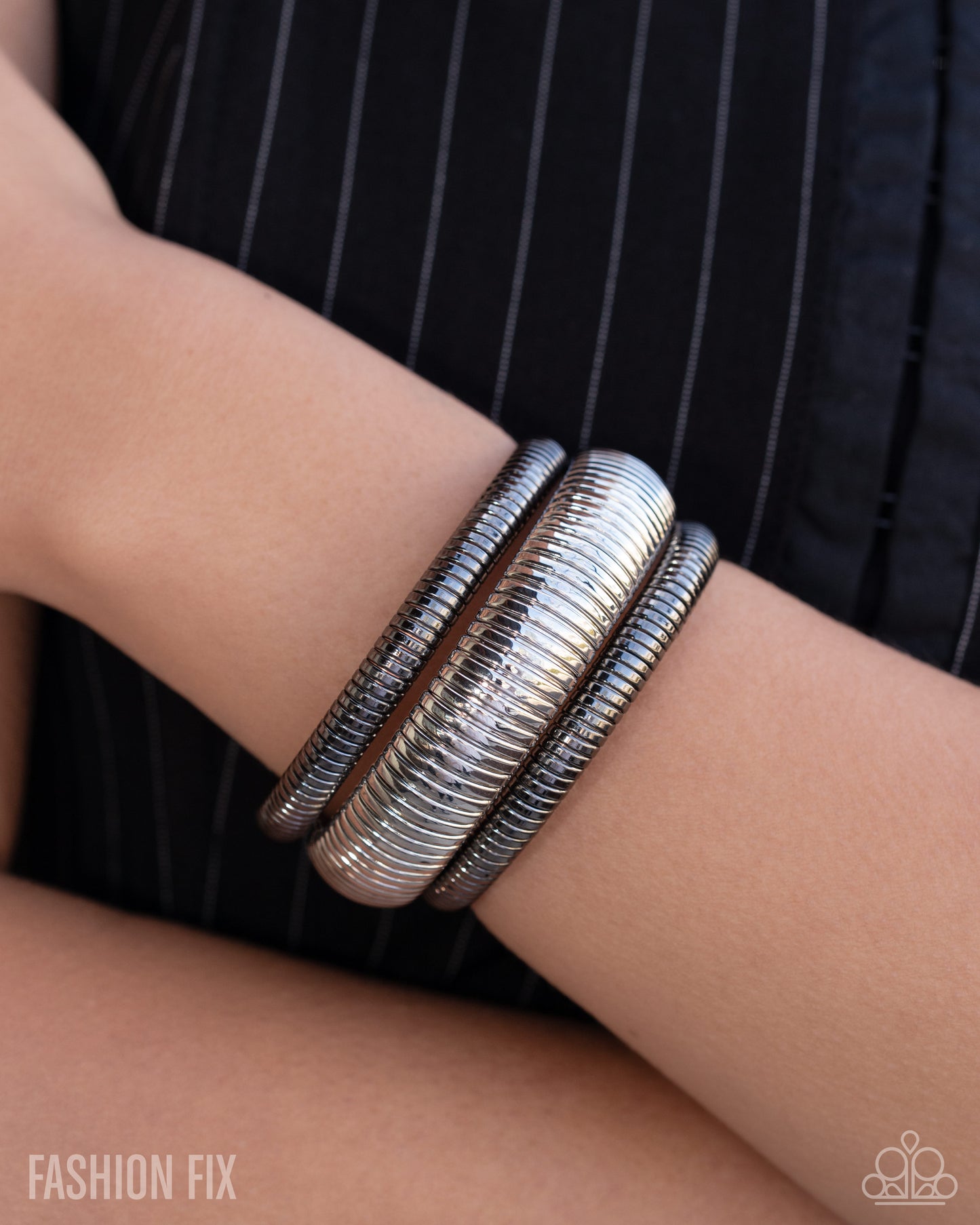 A close-up of a person's wrist wearing three stacked textured silver bangles, with a black pinstriped top in the background. The 'FASHION FIX' logo is visible in the bottom-left corner.