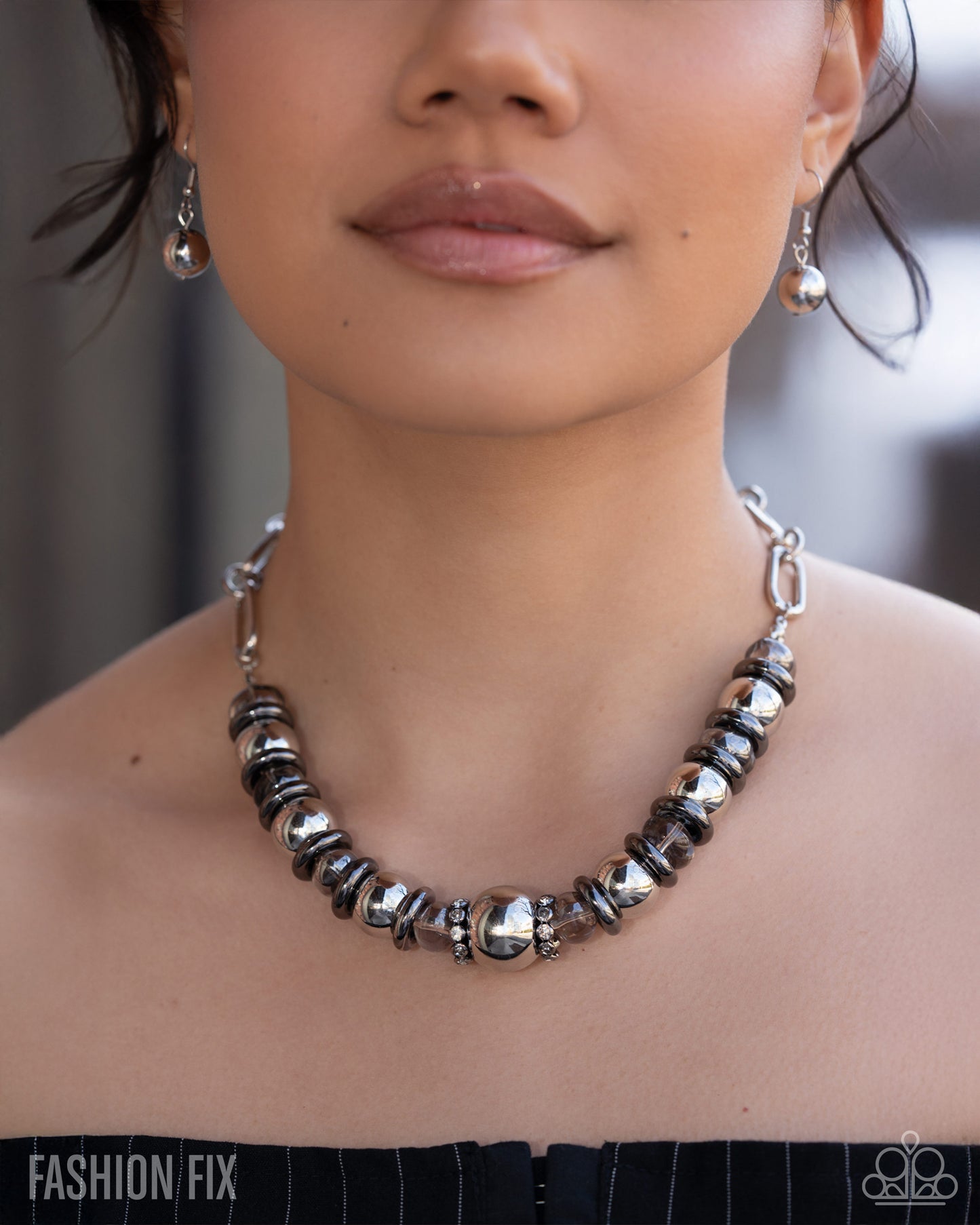 A close-up of a model wearing a chunky statement necklace made of polished silver and black beads, with matching silver dangle earrings. The image has a 'FASHION FIX' watermark at the bottom left.
