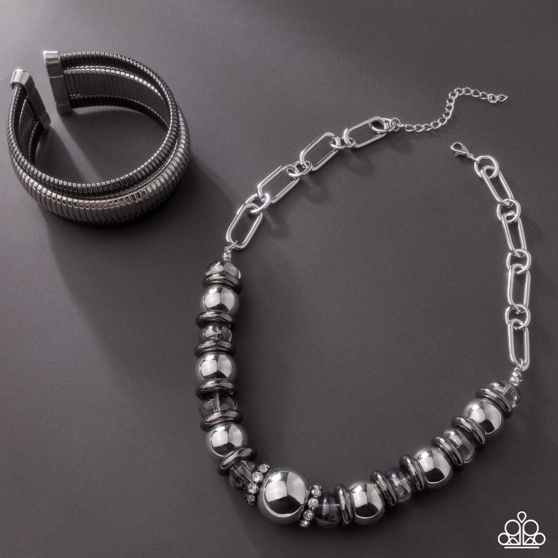 A silver-toned jewelry set featuring a beaded chain necklace and a multi-strand cuff bracelet, displayed on a dark gray background.