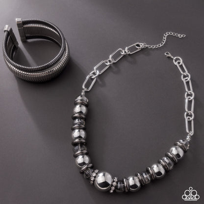 A silver-toned jewelry set featuring a beaded chain necklace and a multi-strand cuff bracelet, displayed on a dark gray background.