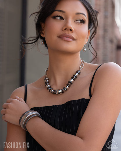 A woman wearing a chunky silver and black beaded statement necklace, silver drop earrings, and a stack of silver bangles, paired with a black pinstripe top. The photo is taken in an outdoor urban setting with a blurred background.
