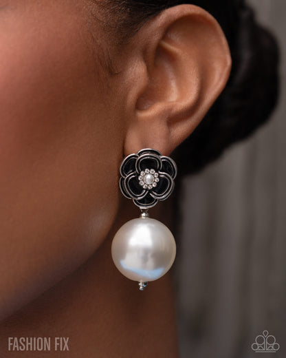 A close-up of a model's ear wearing a dangling earring with a black floral filigree top, a small crystal center, and a large white pearl drop.