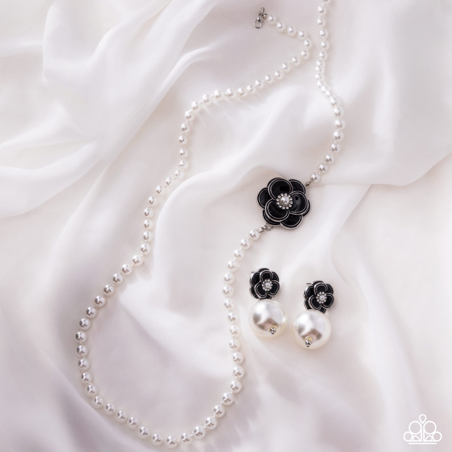 A pearl necklace and matching drop earrings laid on white satin fabric. The jewelry features white pearl beads with black flower-shaped charms and small clear rhinestone centers.