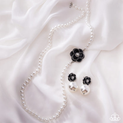 A pearl necklace and matching drop earrings laid on white satin fabric. The jewelry features white pearl beads with black flower-shaped charms and small clear rhinestone centers.