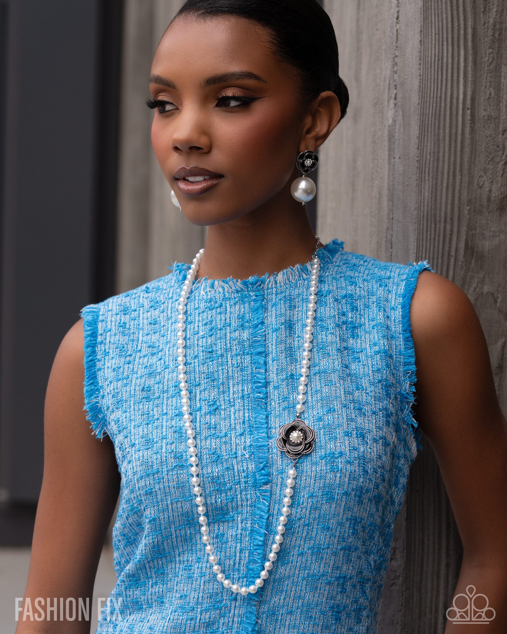 A woman wearing a bright blue textured sleeveless top, a long white pearl necklace with a silver medallion, and matching pearl earrings, posing against a gray wooden wall.