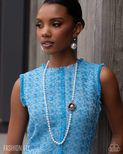 A woman wearing a bright blue textured sleeveless top, a long white pearl necklace with a silver medallion, and matching pearl earrings, posing against a gray wooden wall.