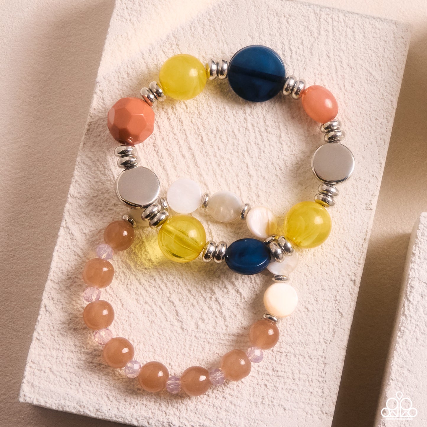Three beaded stretch bracelets displayed on a textured cream card. The bracelets feature translucent orange beads, yellow beads, a dark blue bead, white beads, and silver spacer beads.