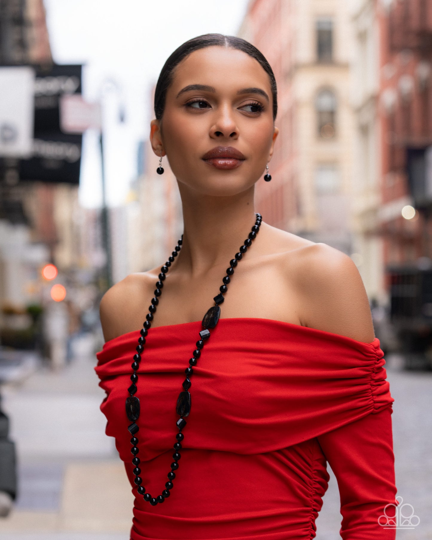 Woman wearing a red off-shoulder dress with a black beaded necklace on a city street.