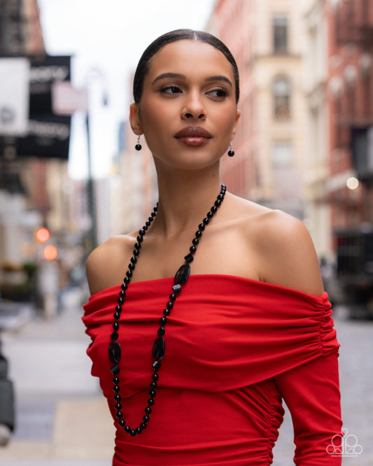 Woman wearing a red off-shoulder dress with a black beaded necklace on a city street.