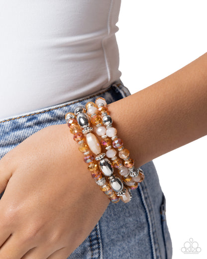 A close-up of a person's wrist wearing three beaded stretch bracelets with orange, white, and metallic silver beads, styled with a white top and blue denim jeans.