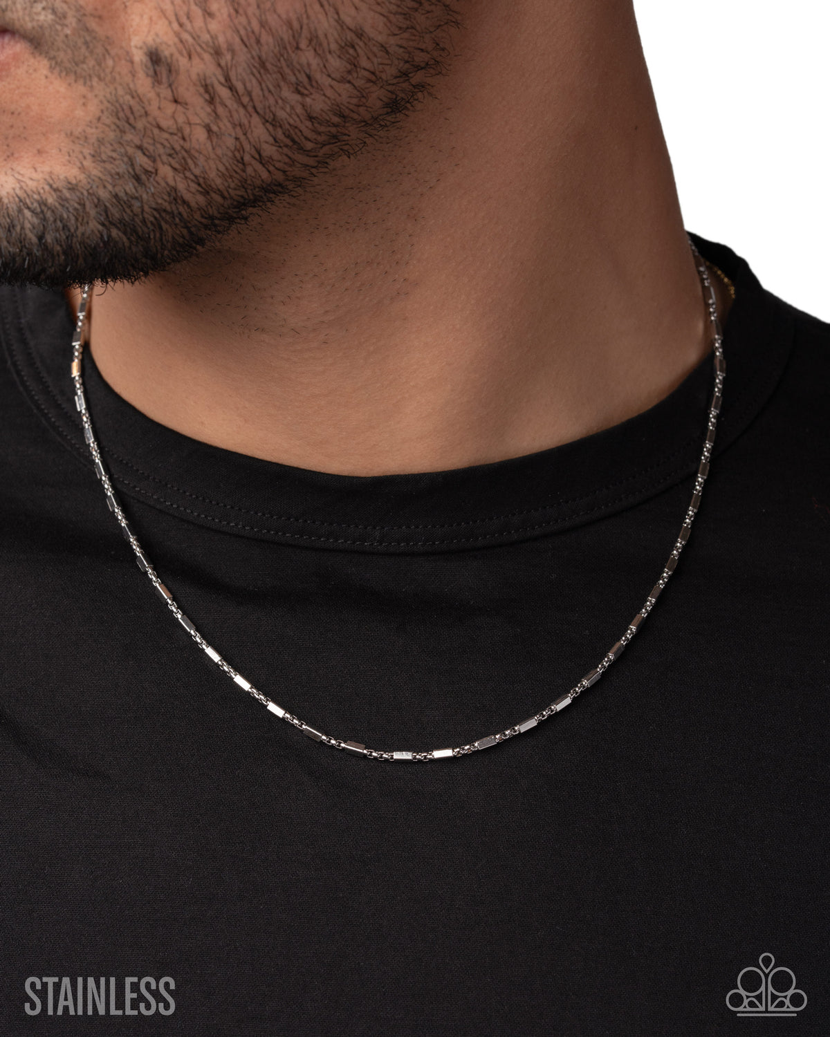 A close-up of a man's neck and lower face wearing a silver stainless steel chain necklace over a black t-shirt. The text 'STAINLESS' is visible in the bottom-left corner, and a brand logo is in the bottom-right.