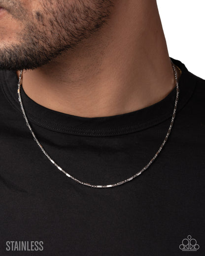 A close-up of a man's neck and lower face wearing a silver stainless steel chain necklace over a black t-shirt. The text 'STAINLESS' is visible in the bottom-left corner, and a brand logo is in the bottom-right.