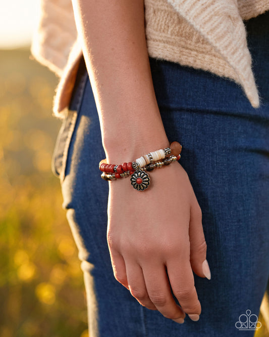 A close-up of a person's hand and wrist wearing a bohemian-style beaded bracelet with red, white, and brown beads, a small sunburst charm, and a bronze-toned metal ring, set against a blurred outdoor background.