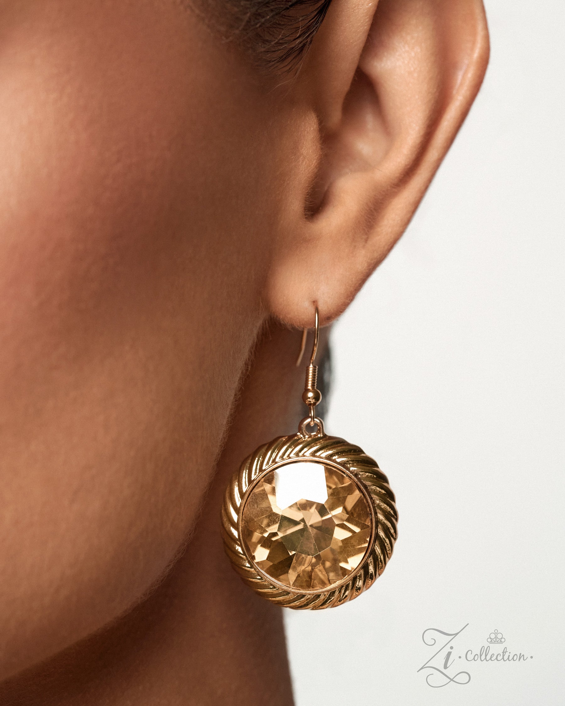 A close-up of a model's ear wearing a gold-toned dangle earring with a faceted amber-tinted round stone and twisted rope-style detailing.