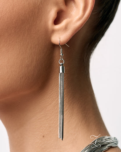 A close-up of a model's ear and neck wearing a long, slender silver dangle earring with a textured metallic finish.