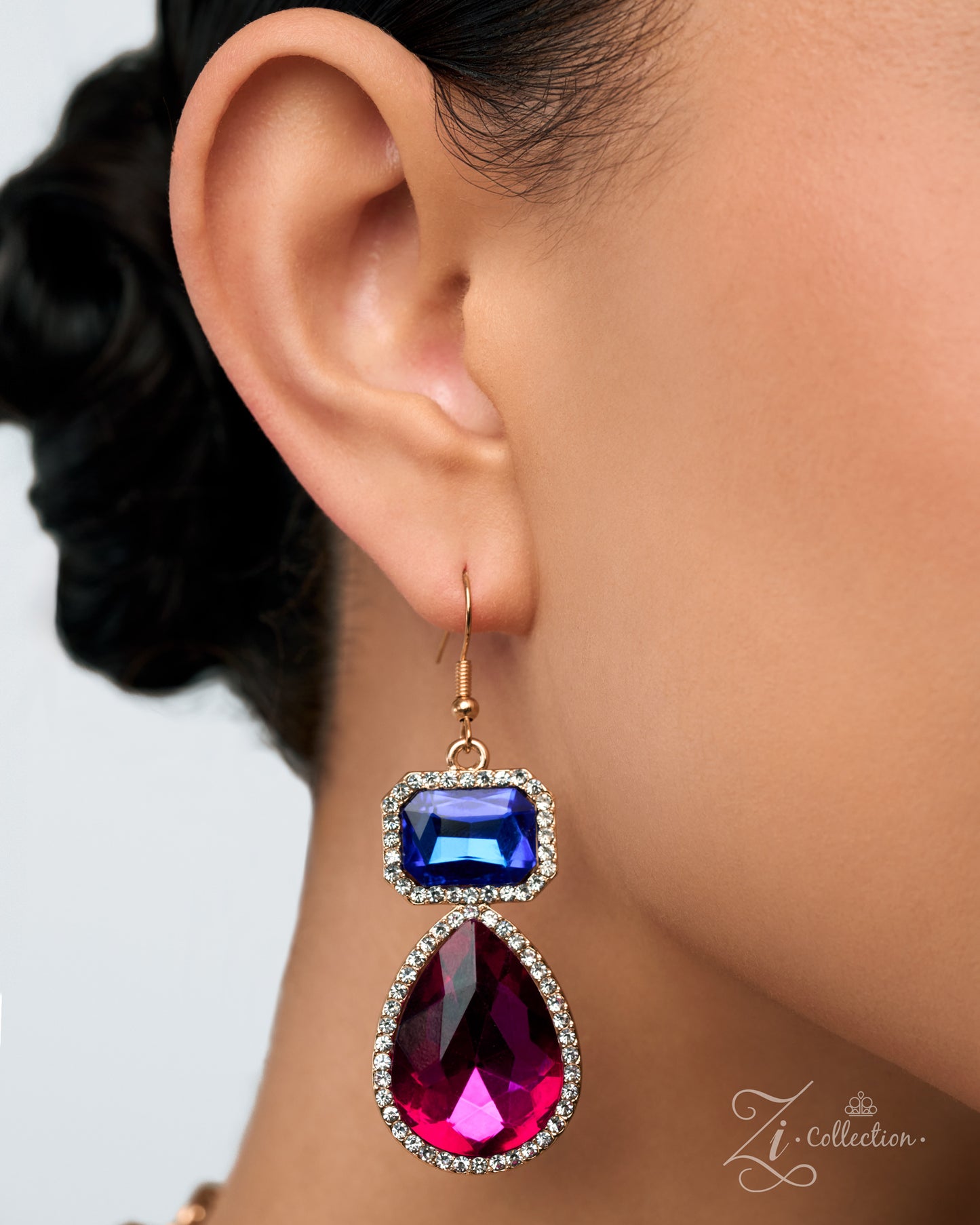 A close-up of a model's ear wearing a gold drop earring with a rectangular blue gemstone and a larger teardrop-shaped magenta pink gemstone, both accented with small clear rhinestones.