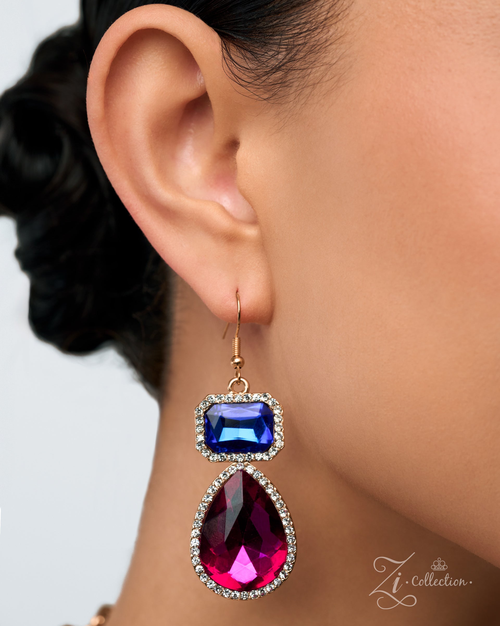 A close-up of a model's ear wearing a gold drop earring with a rectangular blue gemstone and a larger teardrop-shaped magenta pink gemstone, both accented with small clear rhinestones.