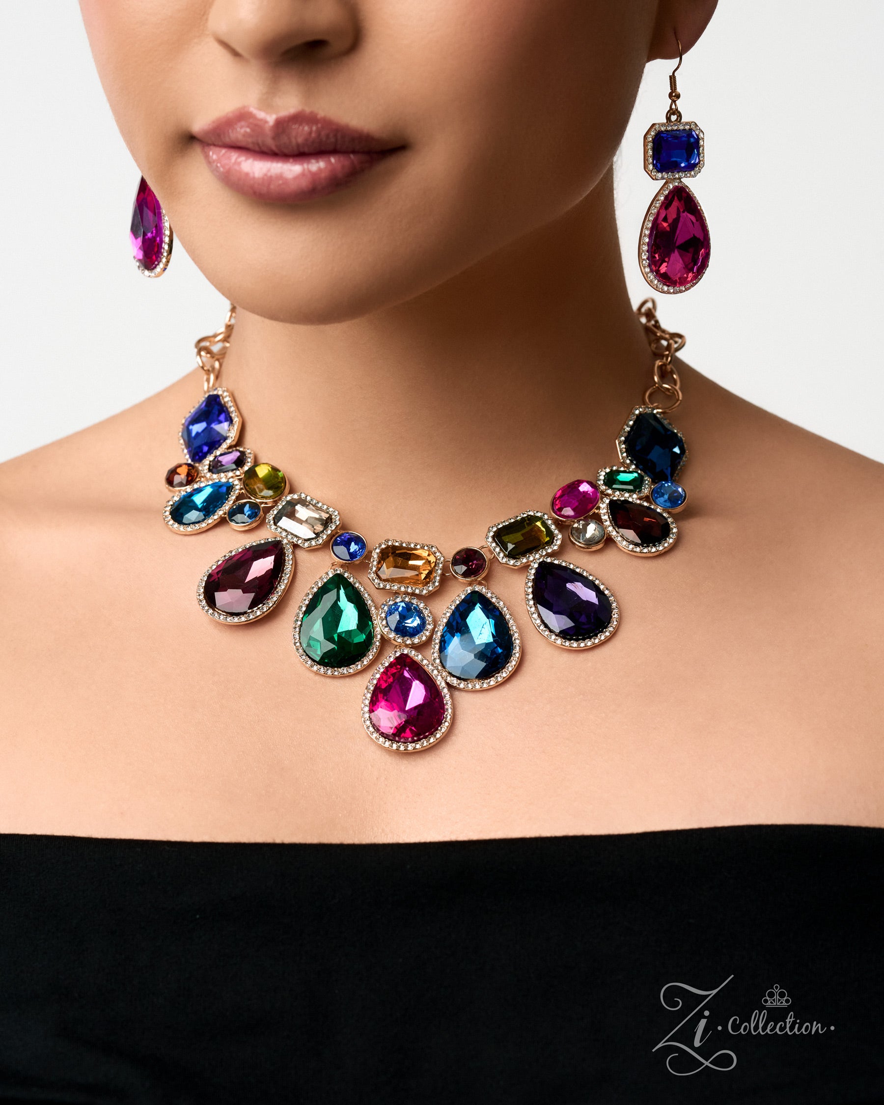 A close-up of a model wearing a gold-plated statement necklace and matching drop earrings with large, colorful teardrop and oval rhinestones in shades of pink, blue, green, and purple, set against a clean white background.