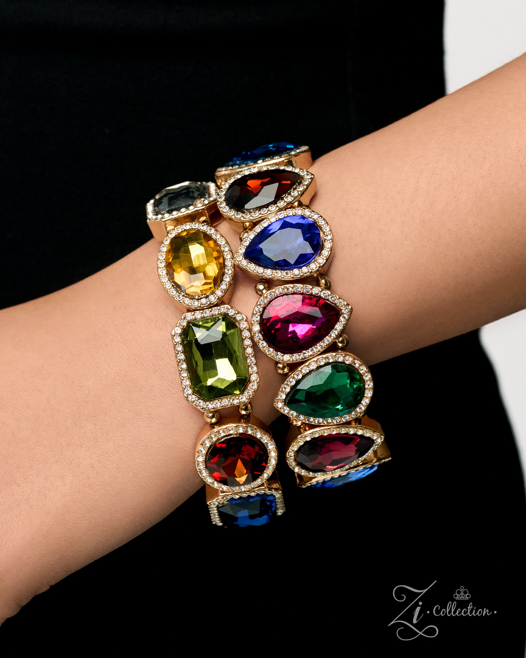 A close-up of a person's wrist wearing a stack of three gold-toned bangle bracelets, each adorned with large, colorful gemstone clusters in shades of blue, green, yellow, red, and purple. The background is a solid black fabric.