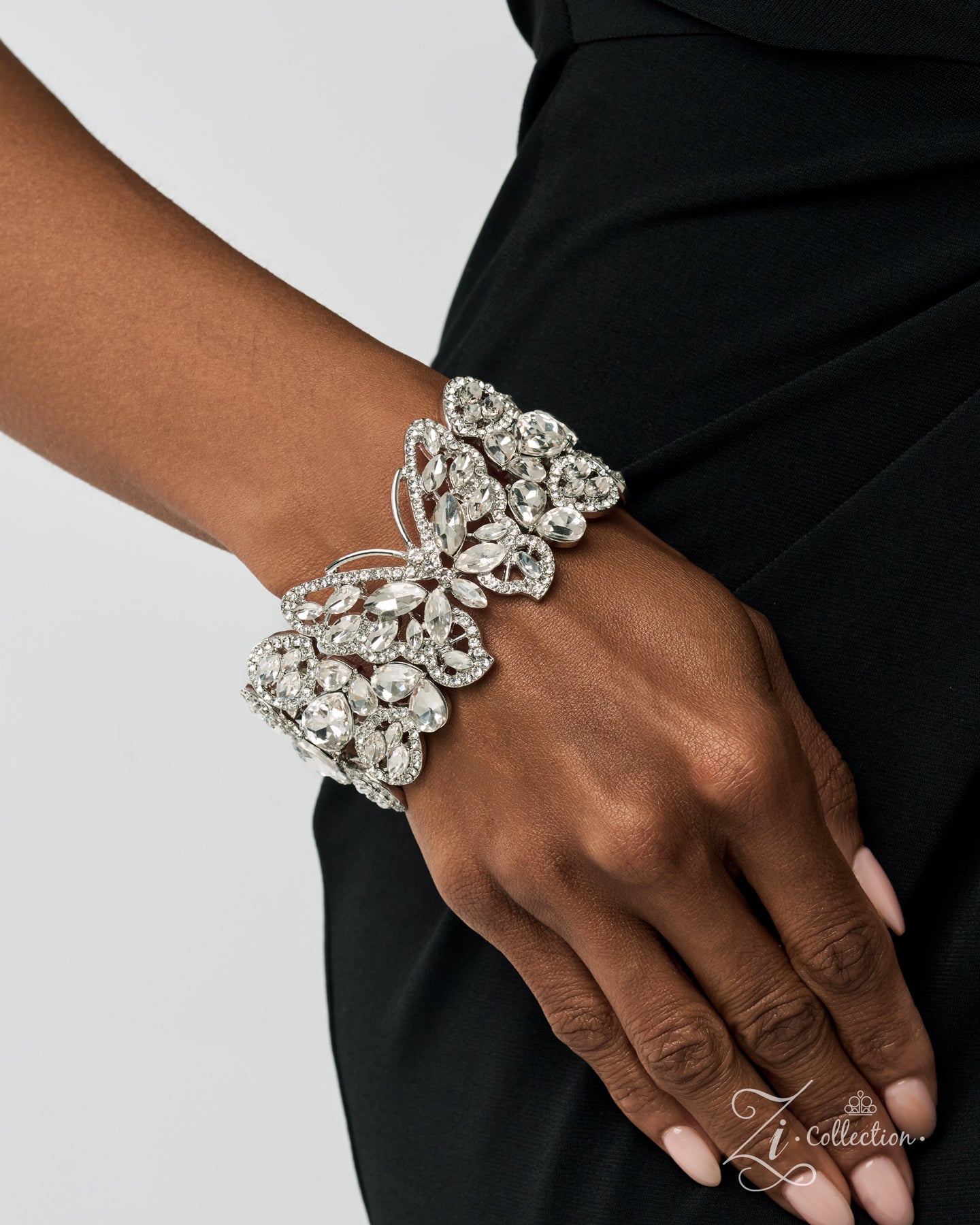 A close-up of a person's hand and wrist wearing a wide, ornate silver-toned cuff bracelet covered in clear crystals, paired with a black dress against a light gray background.