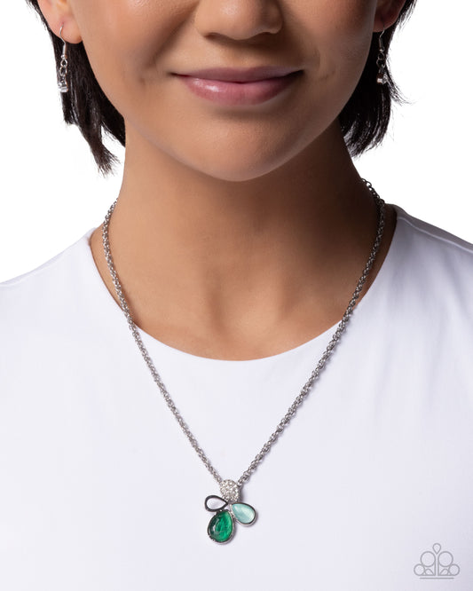 A close-up of a model wearing a silver chain necklace with a bow-shaped pendant featuring a green teardrop gemstone and a light blue oval gemstone, set against a plain white background.
