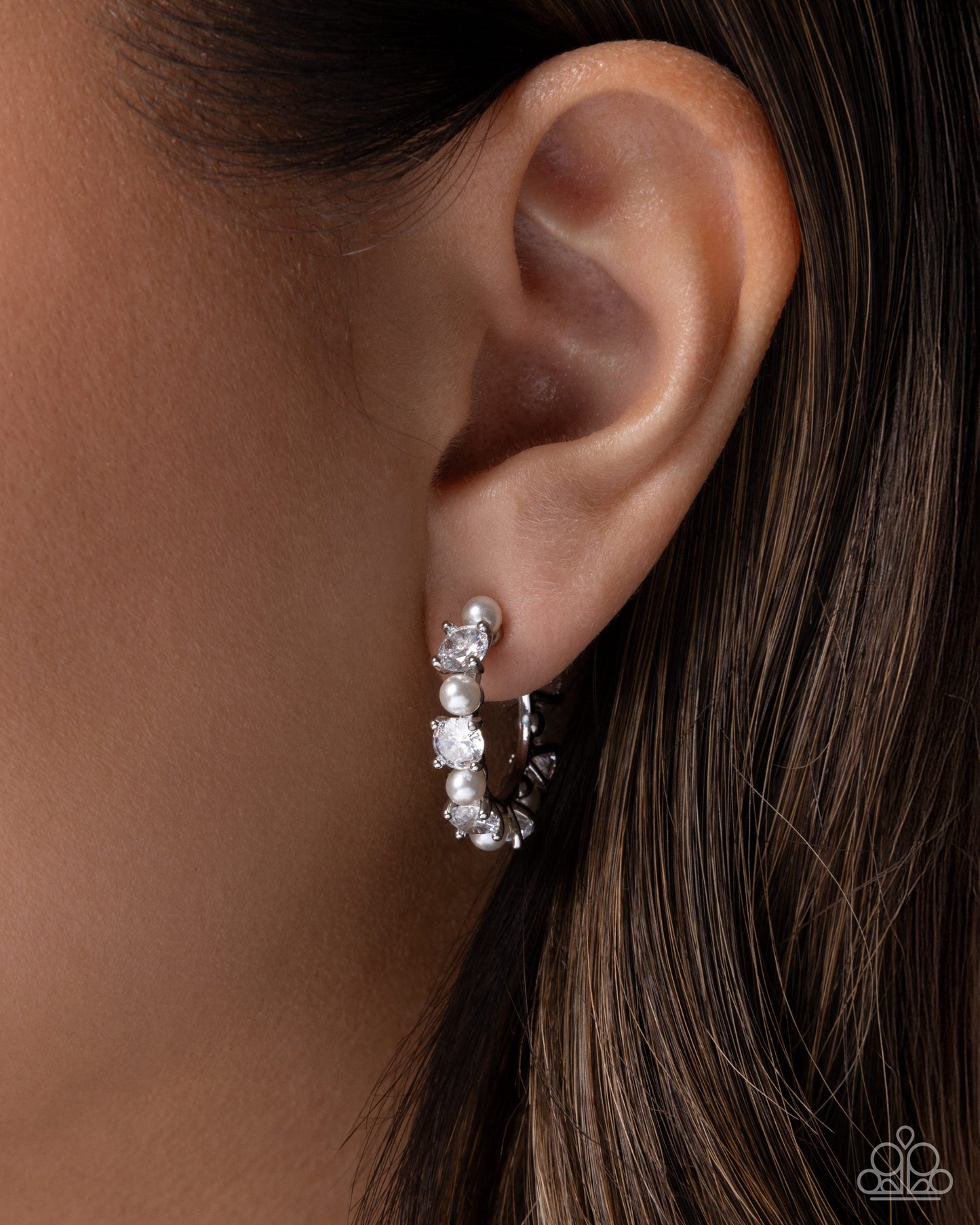 A close-up of a model's ear wearing a silver hoop earring adorned with clear gemstones and a small pearl cluster near the top.