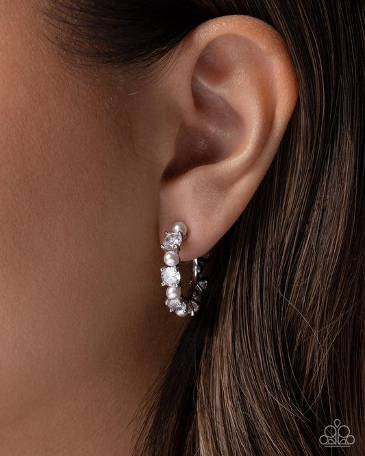 A close-up of a model's ear wearing a silver hoop earring adorned with clear gemstones and a small pearl cluster near the top.