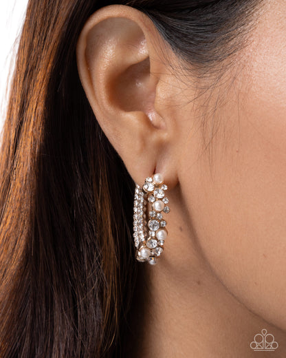 A close-up of a model's ear wearing a pair of silver hoop earrings adorned with clear rhinestones and small white pearls.