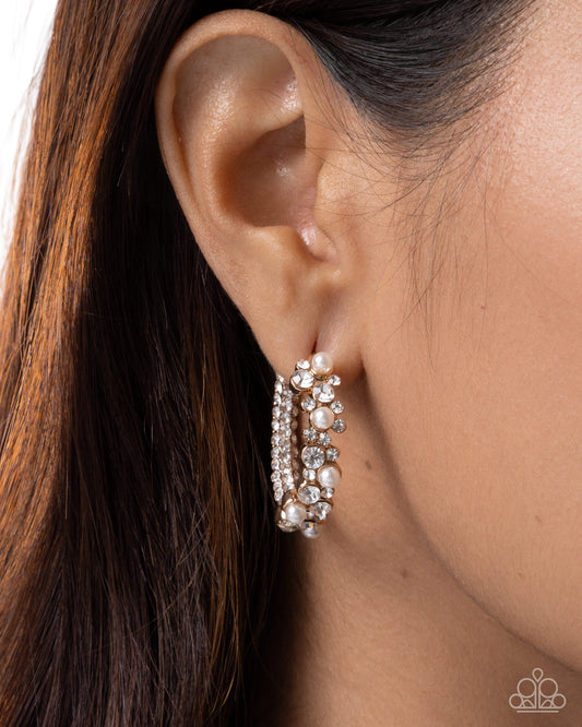 A close-up of a model's ear wearing a pair of silver hoop earrings adorned with clear rhinestones and small white pearls.