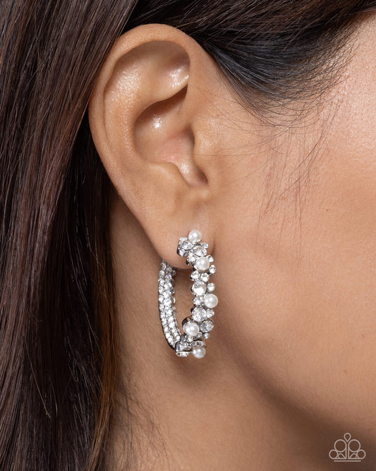 A close-up of a model's ear wearing a silver hoop earring adorned with clear rhinestones and white faux pearls, with dark brown hair framing the side profile.