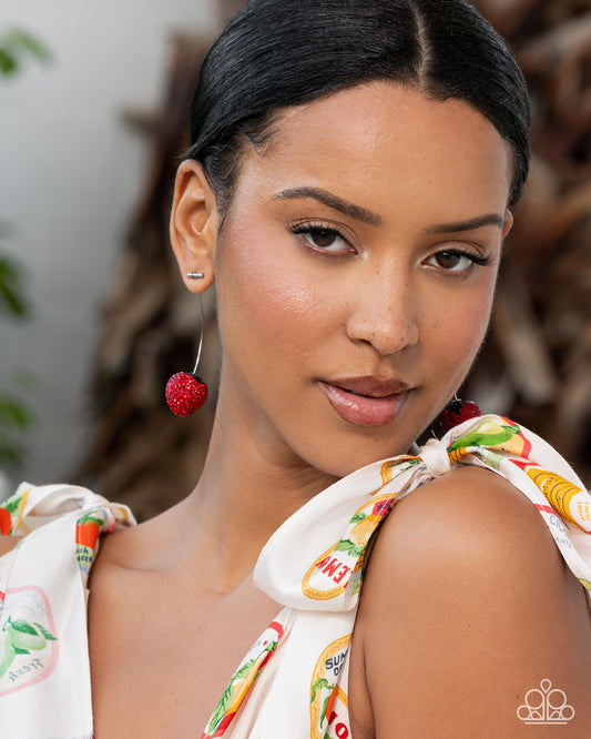 Woman wearing a colorful top with fruit patterns and red earrings.
