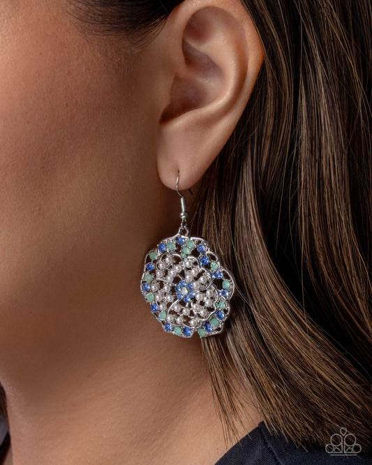 A close-up of a woman's ear wearing a silver-toned dangle earring adorned with clear rhinestones and blue, green, and purple crystals.