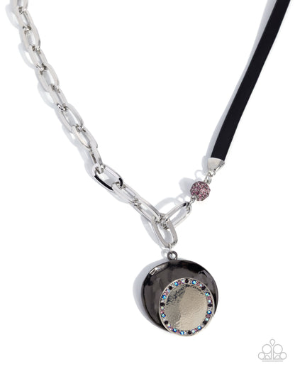A silver-toned chain necklace with a black pendant featuring a textured gold center, a border of rhinestones, and a small pink rhinestone accent on a black cord, shown on a white background.
