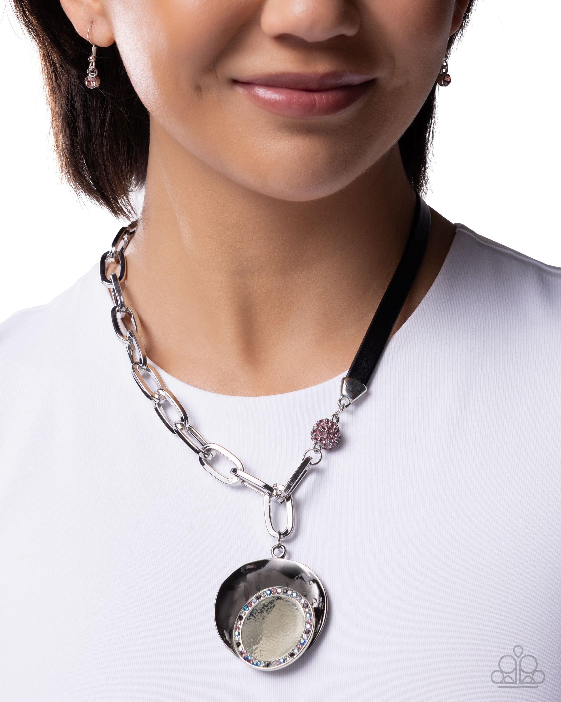 A close-up of a model wearing a silver-toned pendant necklace with a black ribbon strap, a small pink floral accent, and a round silver medallion pendant.